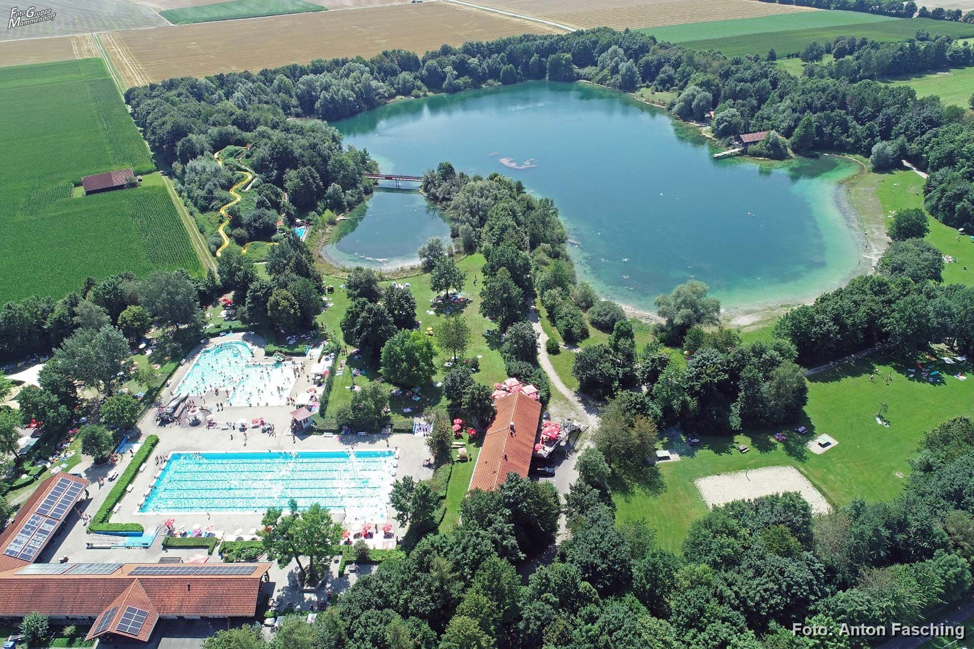 Luftaufnahme des Freizeitpark Mammendorf bei schönem Wetter mit einigen Badegästen. Es ist das Freibad, der Badesee und Liegewiesen zu sehen. Im Hintergrund sind weite Getreidefelder zu sehen.