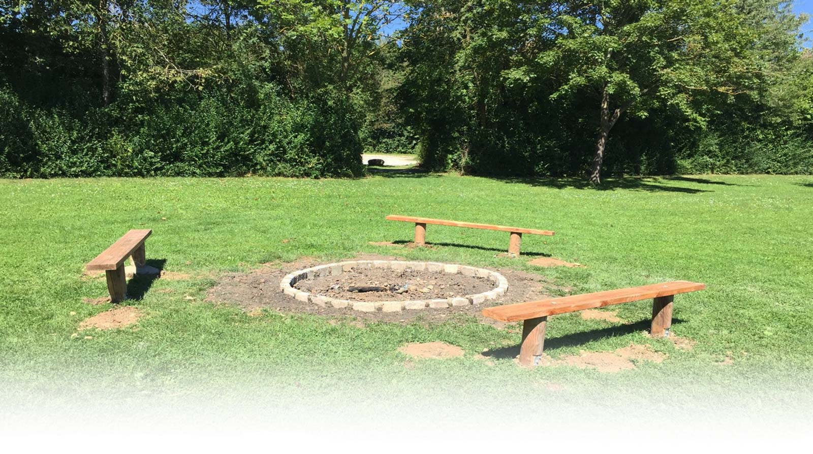 Große runde Grillstelle auf der Liegefläche des Badesees Mammendorf bei schönem Wetter. Grüne Wiese und grüne Bäume.