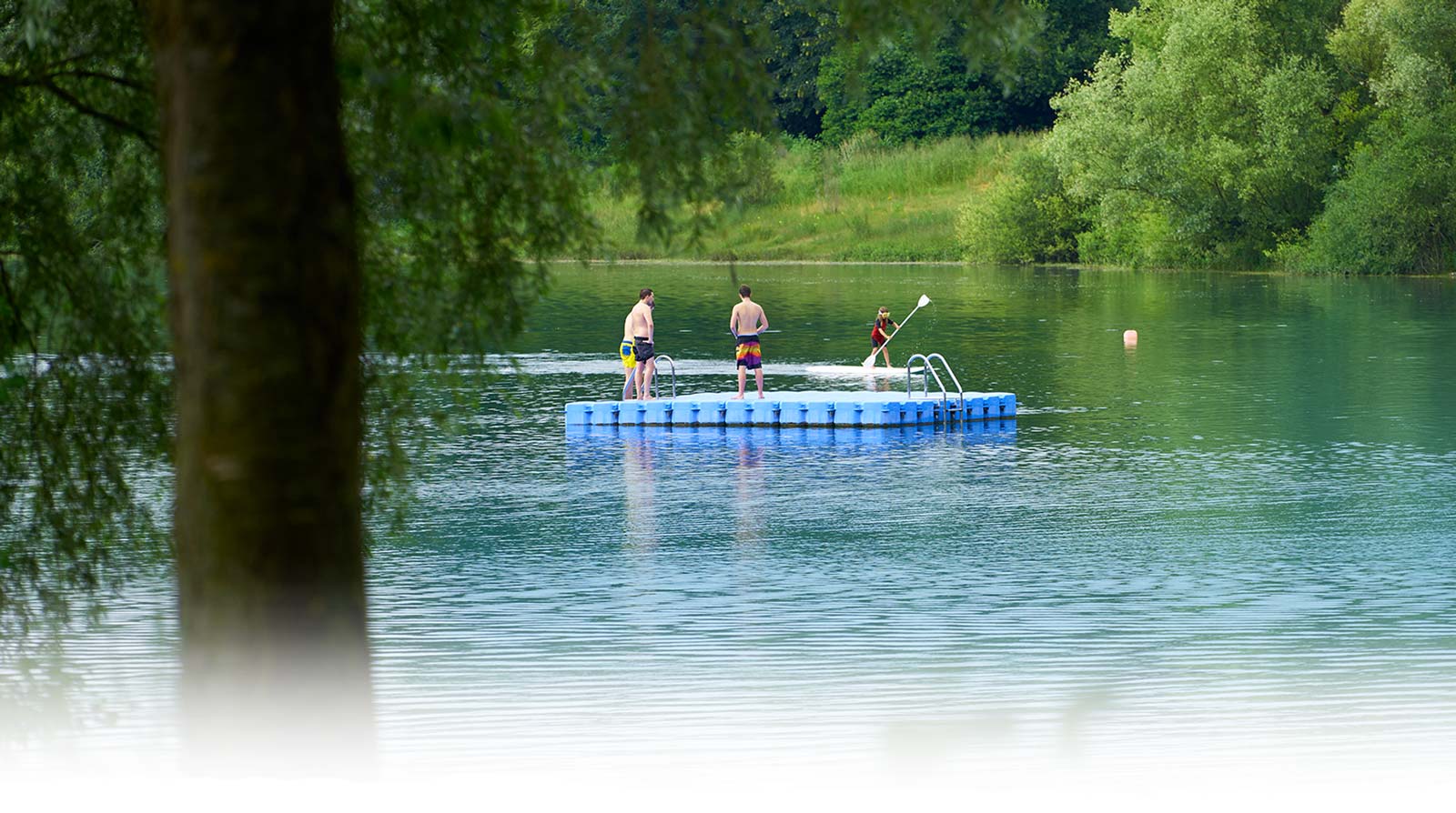 Drei junge Männer auf blauer Schwimminsel im Badesee Mammendorf. Im Hintergrund sieht man ein Kind mit Taucherbrille auf einem Sub mit einem Paddel.