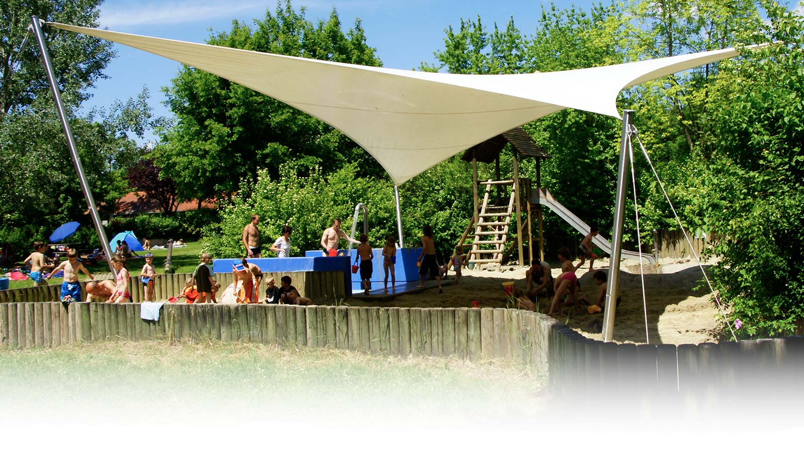 Sandspielplatz mit beige-farbenen Sonnensegel des Freibads Mammendorf. Kleinkinder spielen im Sand mit ihren Eltern.