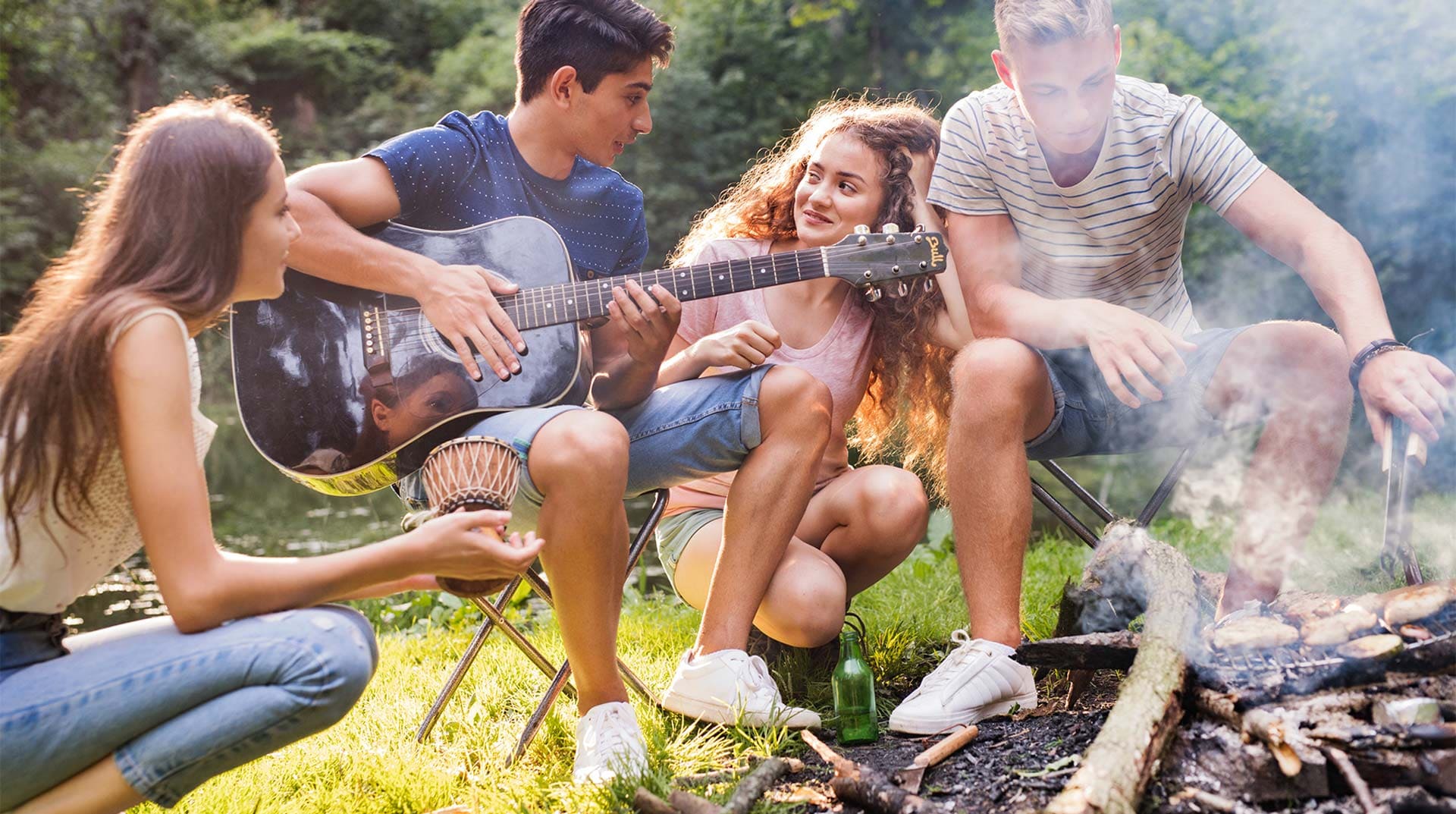 Vier Jugendliche sitzen gemeinsam an einer Grillstelle, ein Junge spielt Gitarre für ein Mädchen, ein Junge wendet das Fleisch und ein Mädchen schaut zu