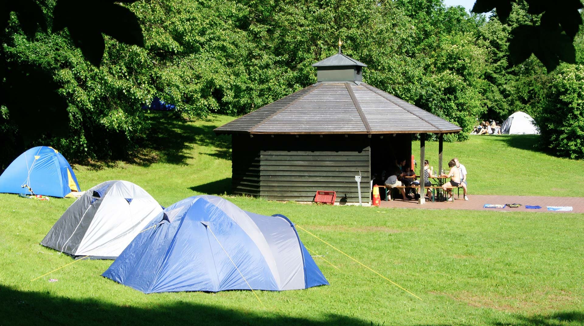 Holzpavillion des Zeltplatz Mammendorf in der Mitte der grünen Liegewiese umringt von kleinen blauen Zelten. Zeltplatzbesucher sitzen vor dem Pavillion auf einer Bierbank und essen.